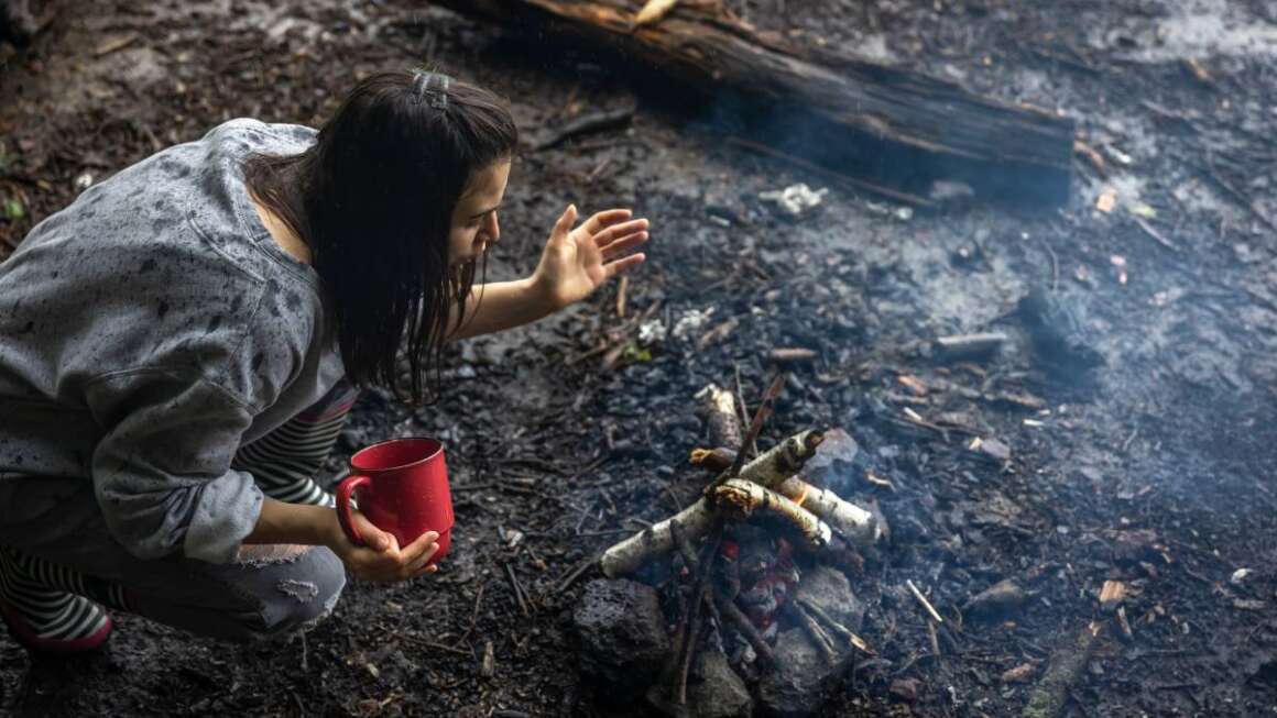 A girl with a cup in her hand is fanning the fire to keep warm.