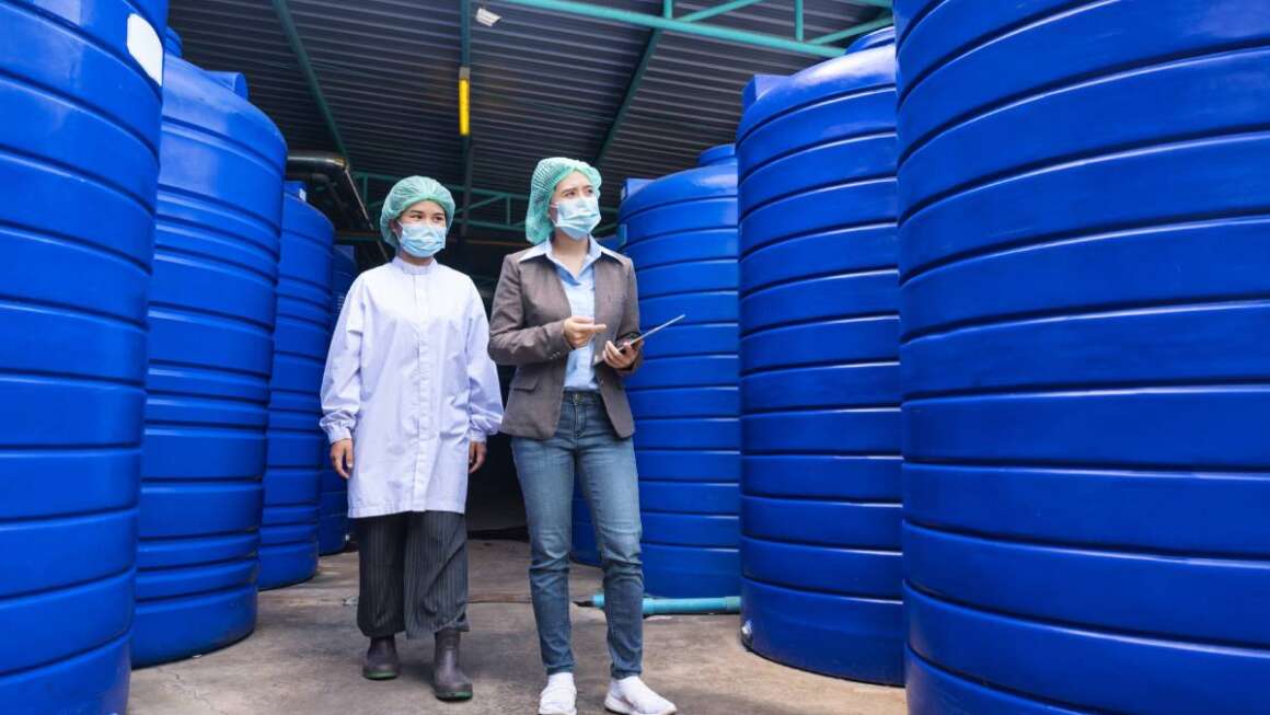 Asian inspector supervisor woman and assistant employee wearing face mask to checking stock of water tank in storage area at beverage processing plant